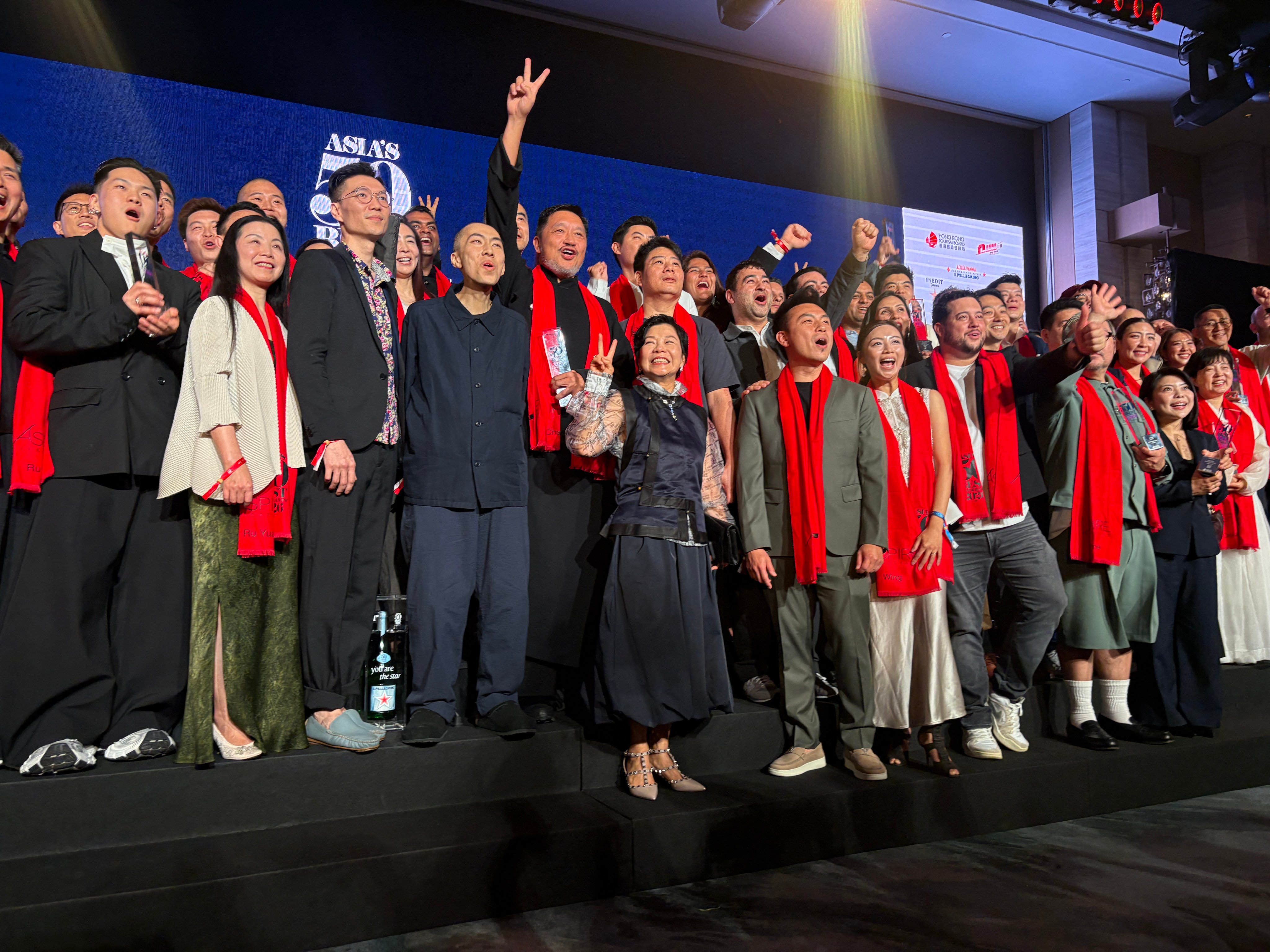 Large group of people on stage wearing red scarves at Asia's 50 Best Restaurants 2026 ceremony, blue backdrop visible behind them.