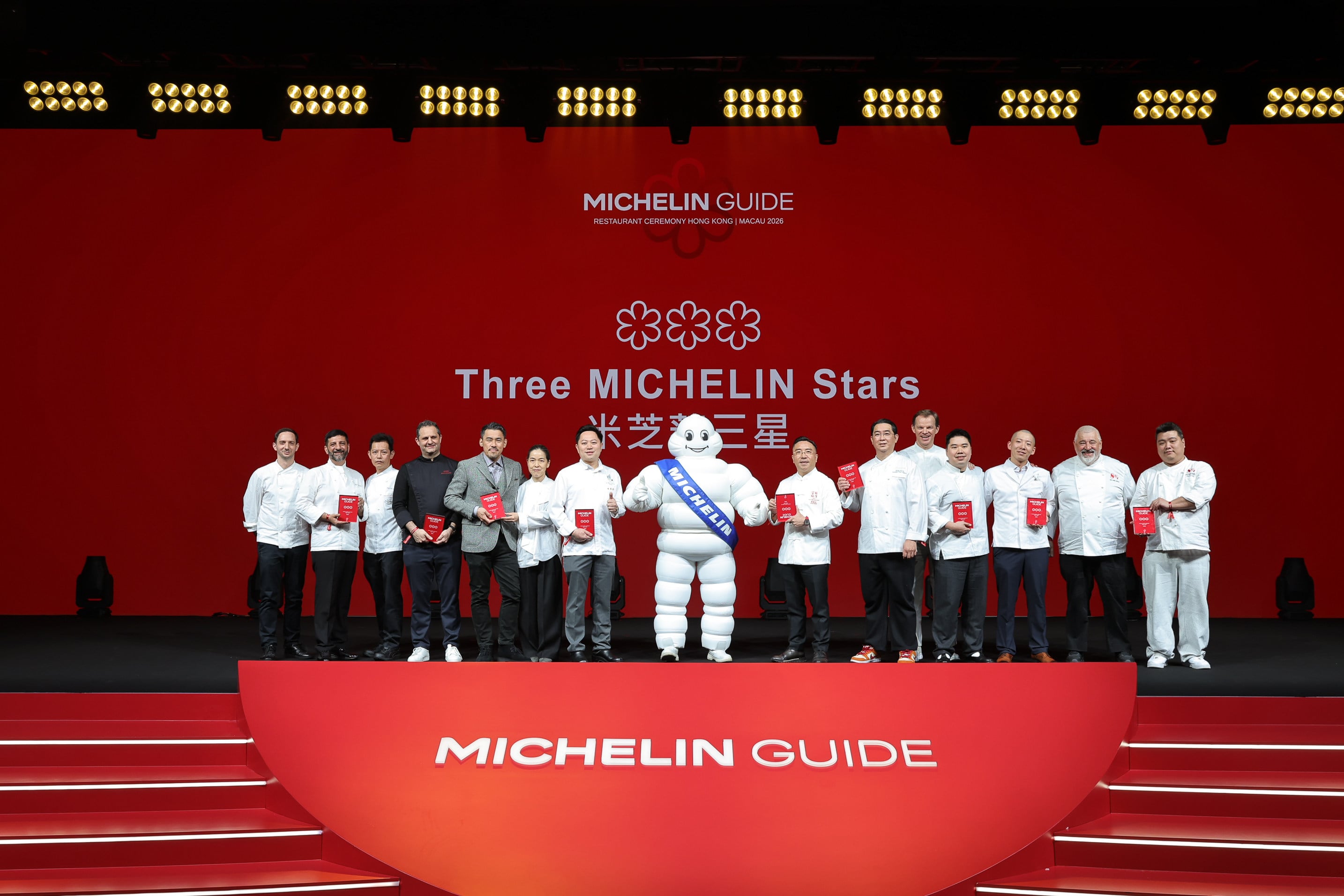 MICHELIN GUIDE ceremony stage with chefs in white coats holding red award certificates, Michelin mascot, and three-star symbol backdrop at Hong Kong M
