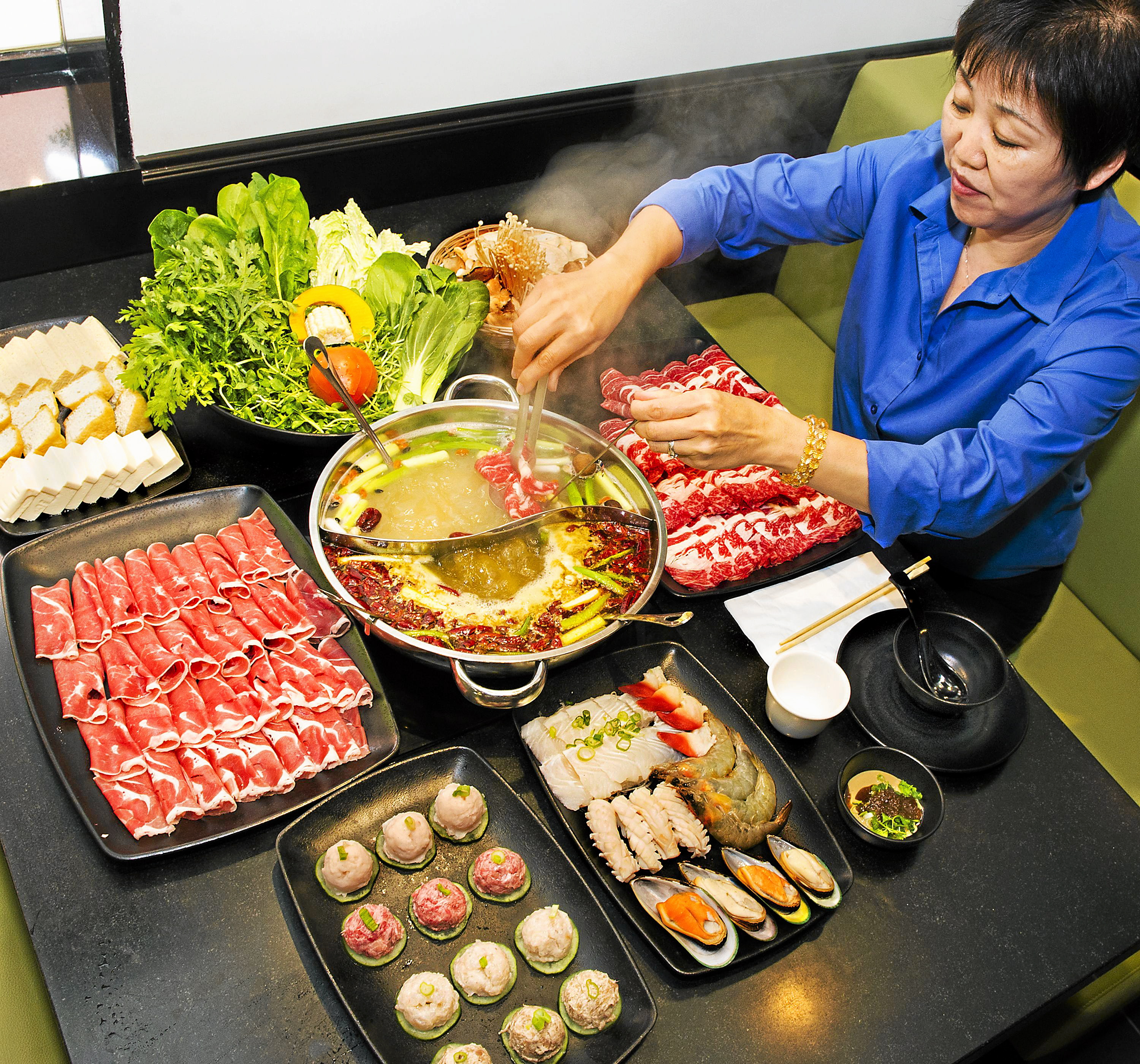 Hot pot dining table with sliced raw beef, seafood, vegetables, and broth in divided pan; server in blue shirt adding noodles with chopsticks.