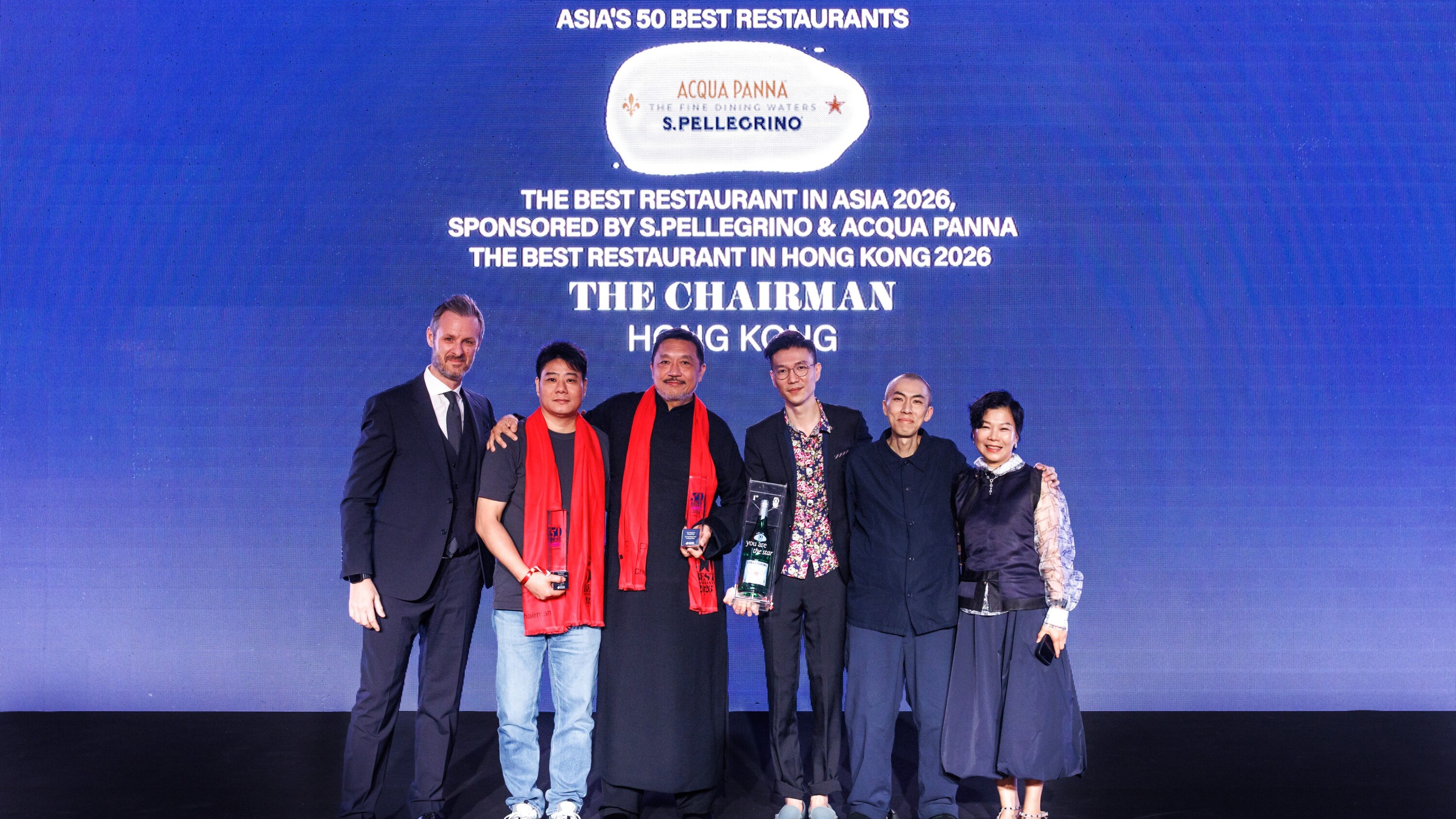 Award ceremony stage with six people holding trophies, blue backdrop displaying 'ASIA'S 50 BEST RESTAURANTS' and 'ACQUA PANNA S.PELLEGRINO' branding, 