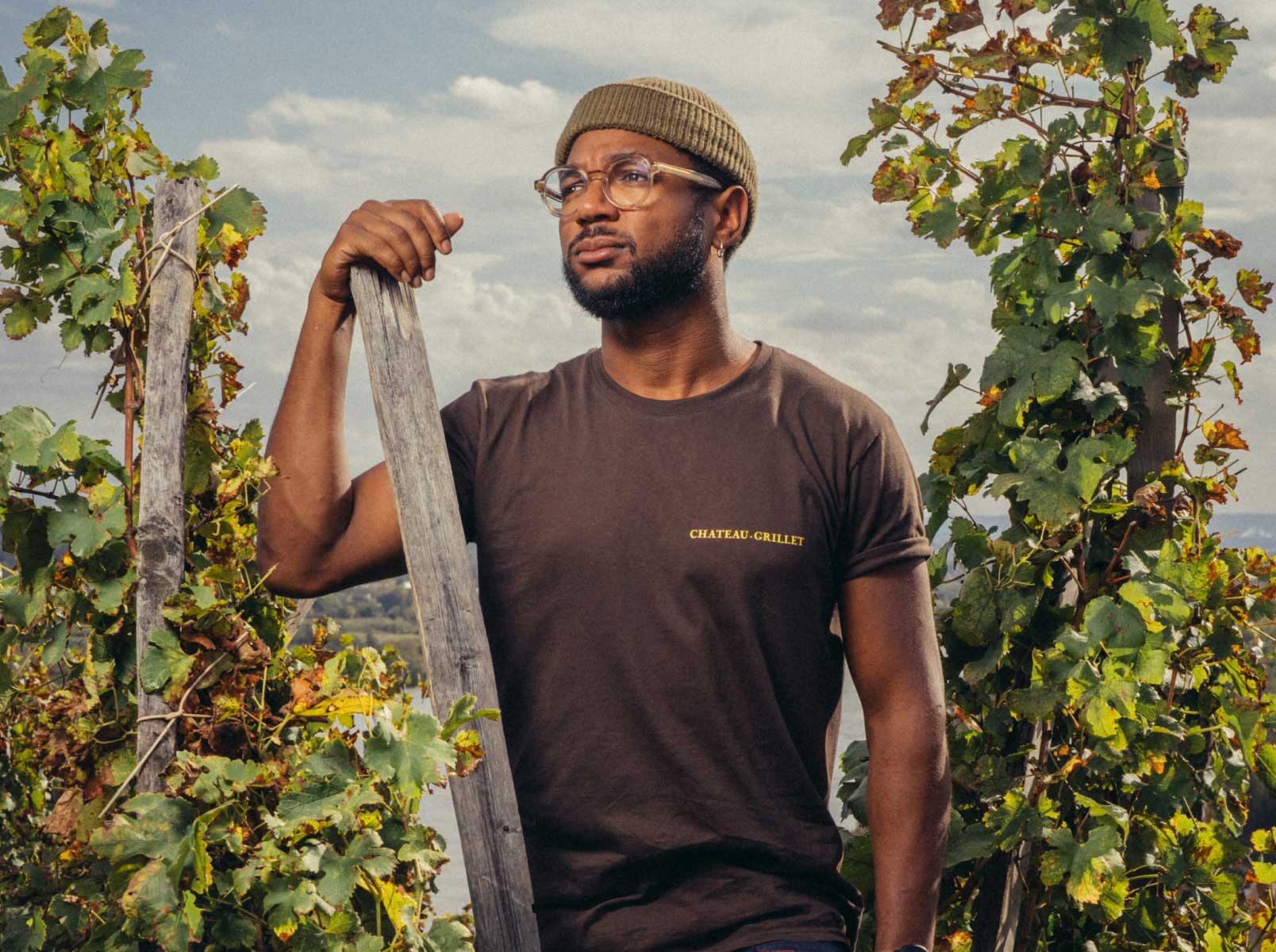 Man in CHATEAU-GRILLET t-shirt holding wooden stake among vineyard rows, wearing beanie and glasses