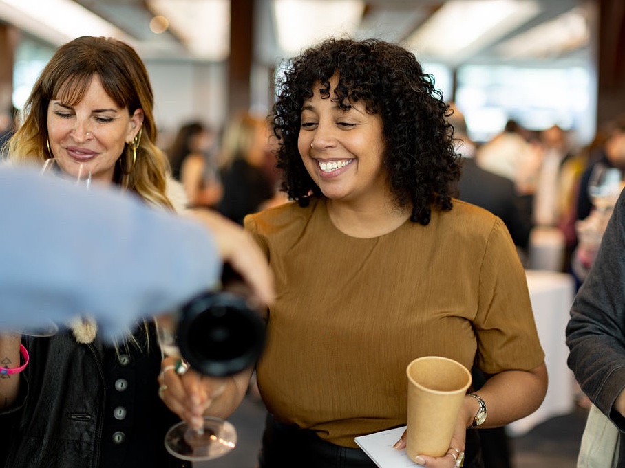 Two women smiling at a social gathering, one wearing a tan shirt holding a cup, another in a light blue top, with other attendees visible in the background of a bright indoor space.