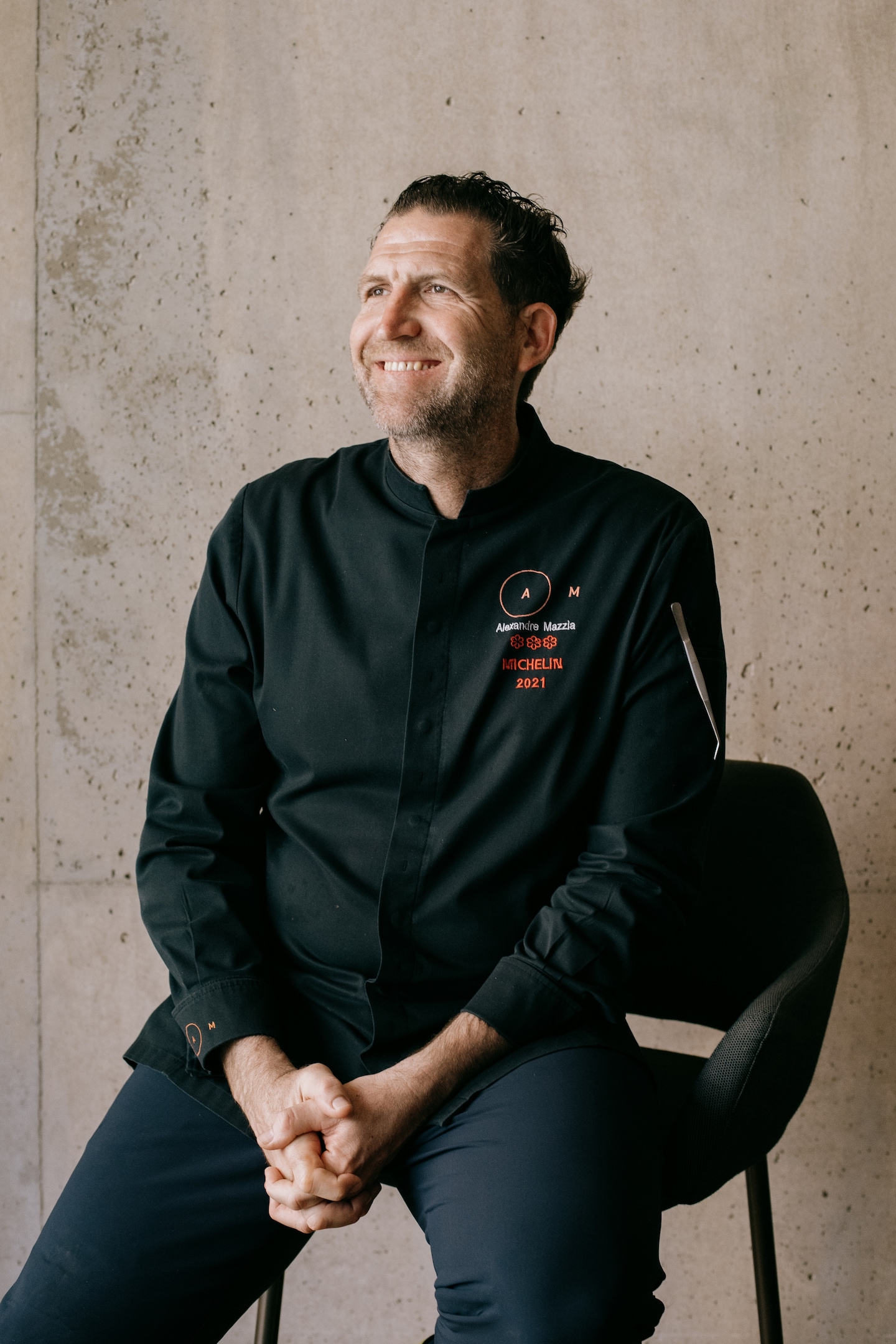 Chef in black chef's coat with 'Alexandre Mazzla' and 'MICHELIN 2021' embroidered in red, smiling against textured concrete wall.