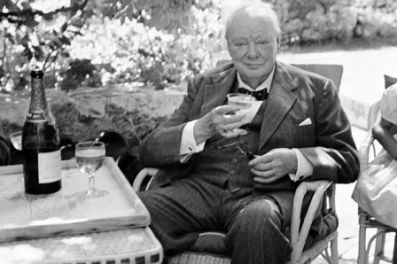 Winston Churchill seated outdoors with a Champagne glass, a bottle and table beside him.