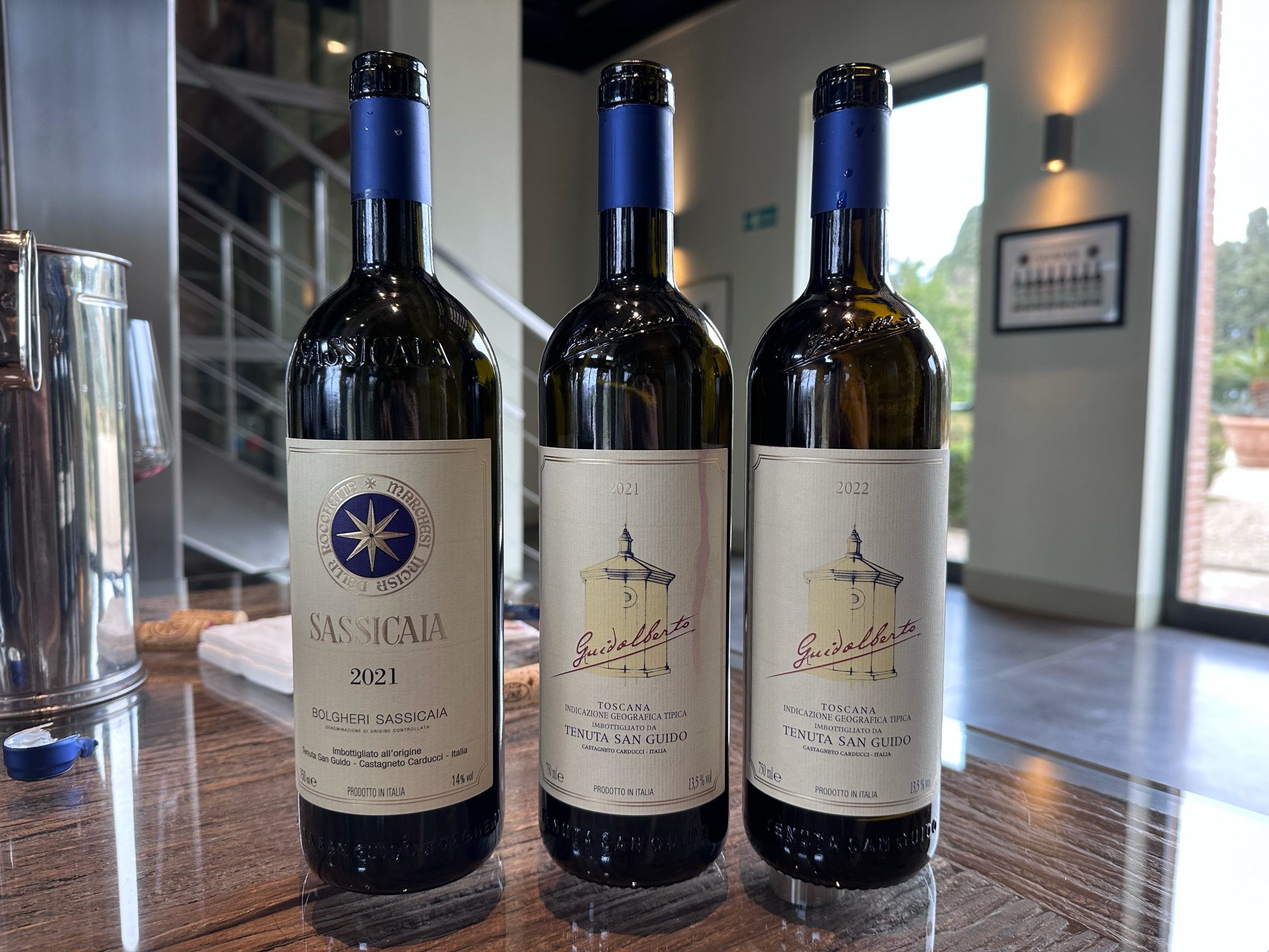 Three wine bottles with blue capsules displayed on a wooden table: Sassicaia 2021, Guidealberta 2021 Toscana, and Guidealberta 2022 Toscana.